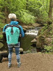 lady with a small black dog wearing the mint green coloured waterproof bag . She is in a  woodland area looking down at a river with a small waterfall