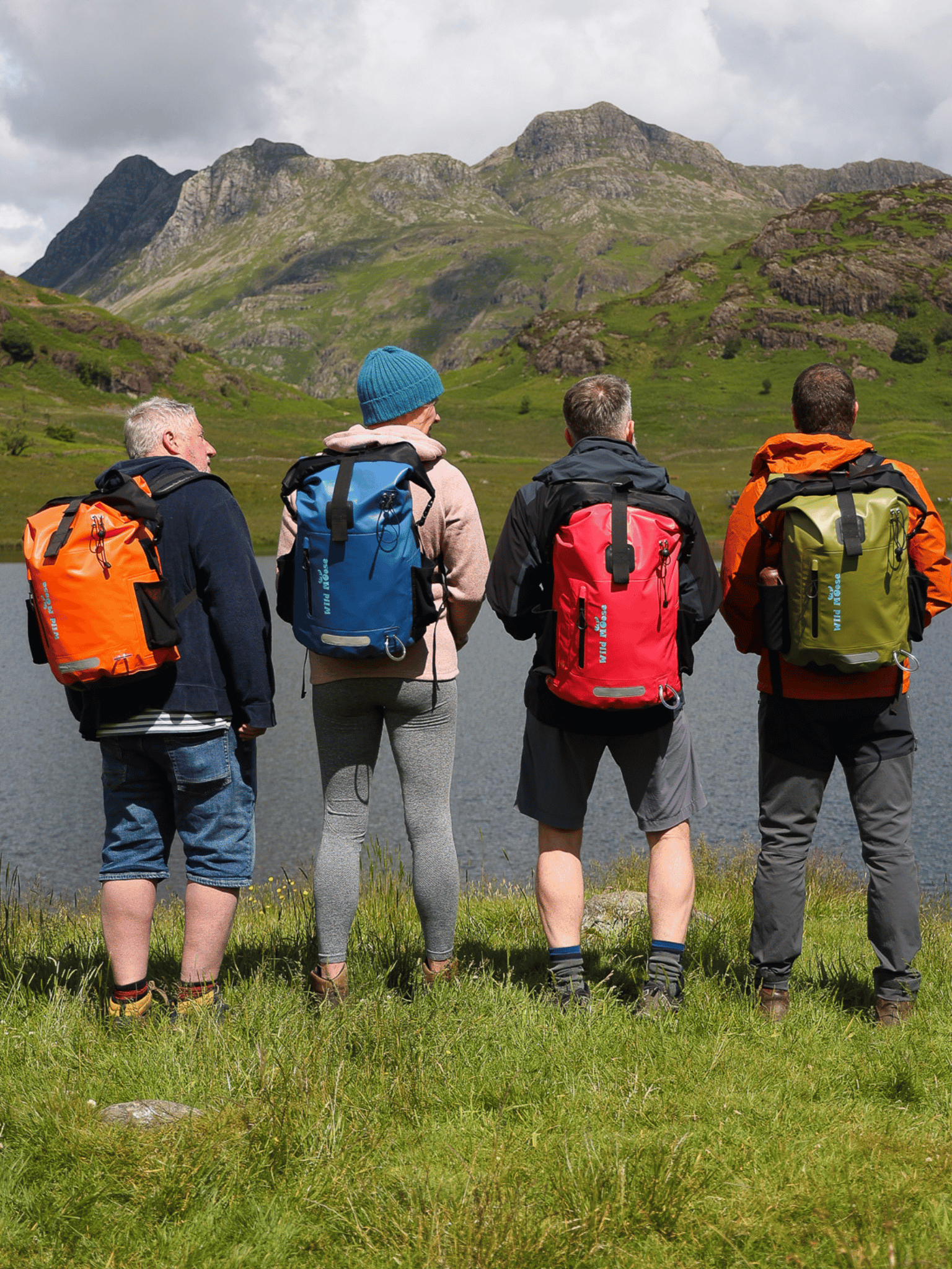4 people stood in front of Blea Tarn in Little Langdale, each wearing a different colour backpack. Flame orange, electric blue, hot pink and olive green