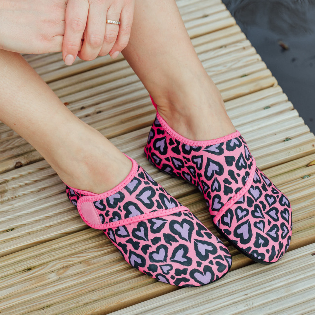 Pink and black leopard print neoprene beach shoes worn by a person sitting on a wooden deck.