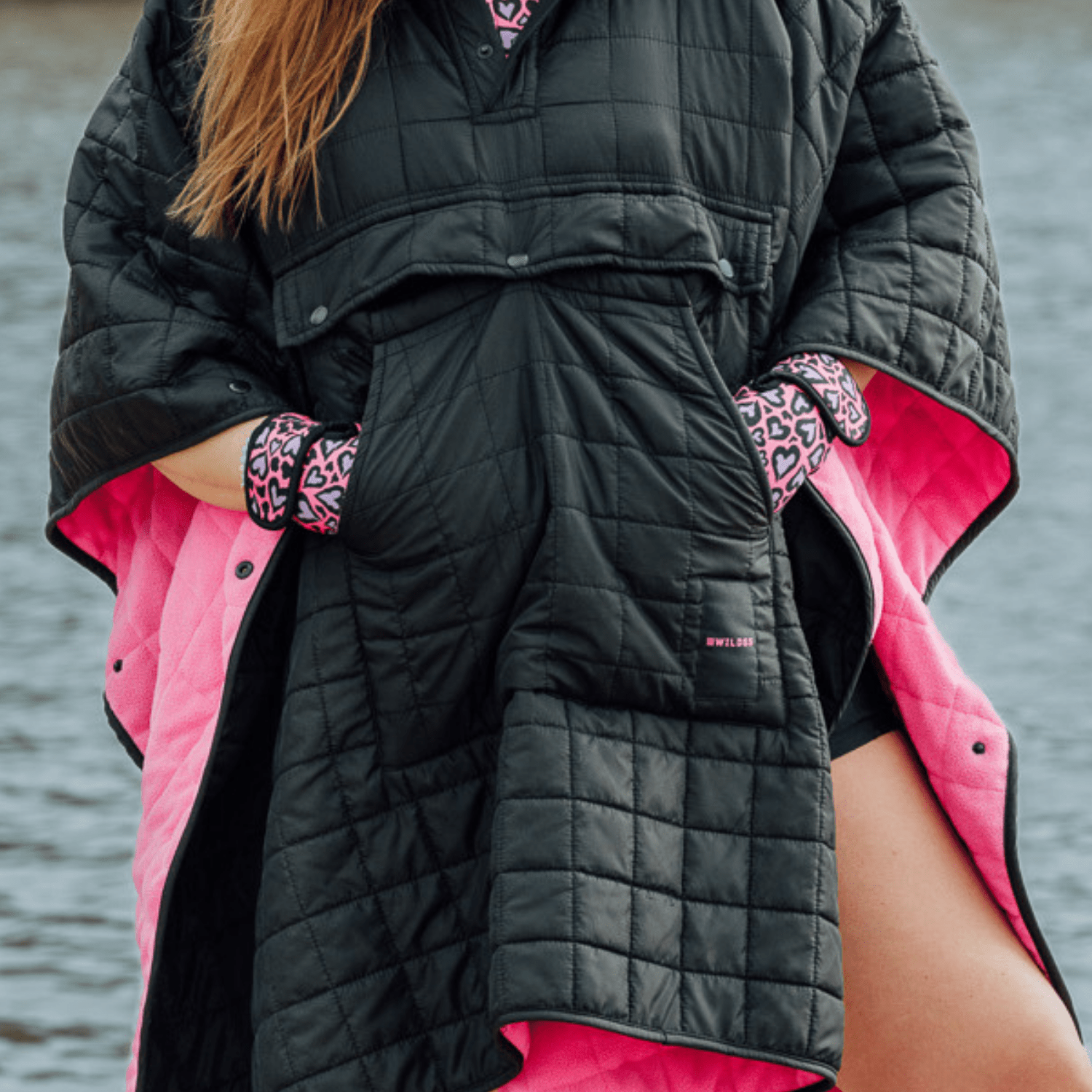 Image of woman wearing weatherproof black robe with large front pocket, side popper fastening, hood and pink fleece lining