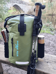 an olive green waterproof backpack on a bench in front of a bridge going over a rive. there are walking poles attached the backpack and a mat and a flask in the side pockets