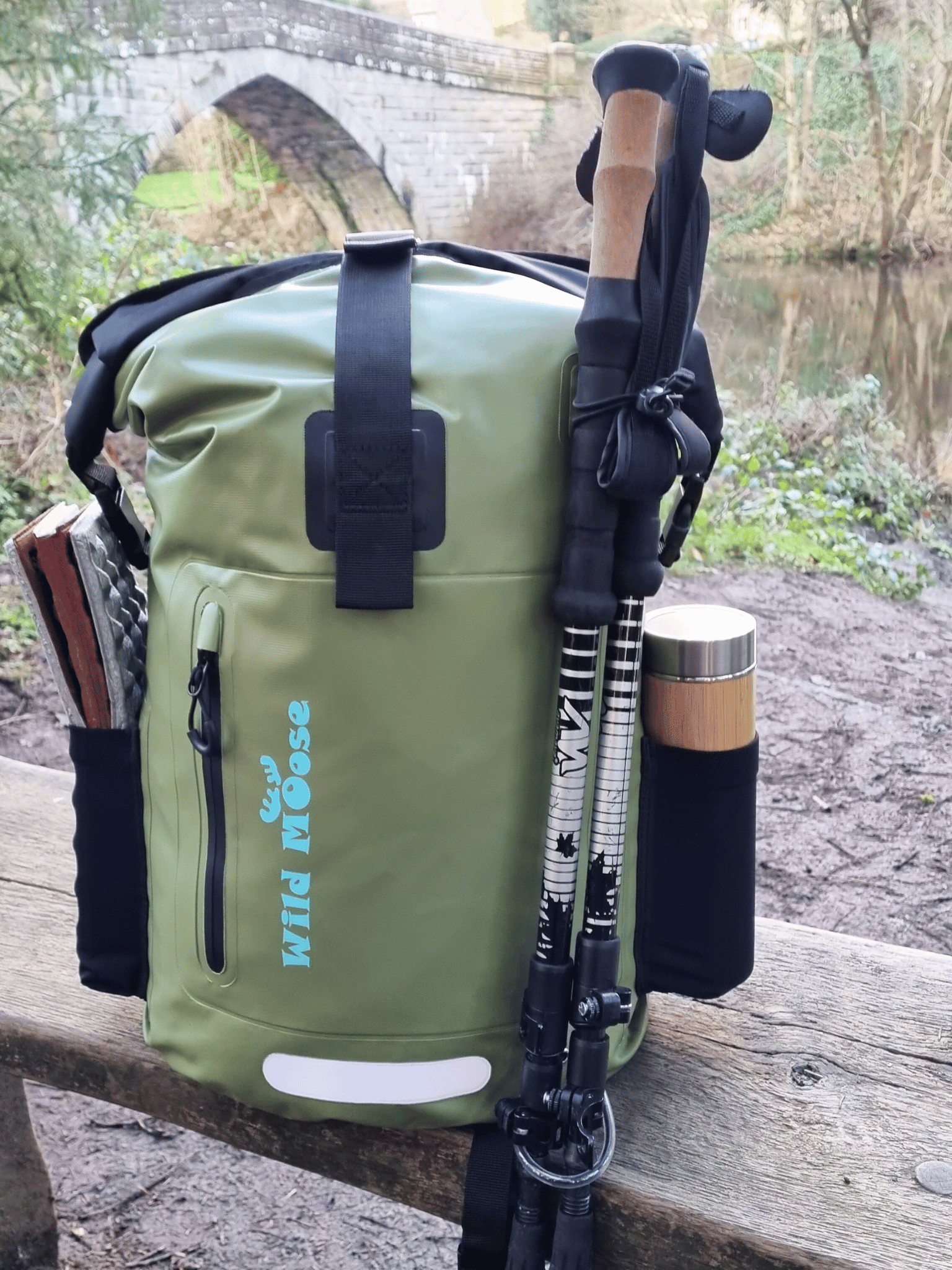 an olive green waterproof backpack on a bench in front of a bridge going over a rive. there are walking poles attached the backpack and a mat and a flask in the side pockets