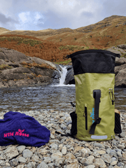 a green backpack and a blue Wild moose poncho towel on a pebble beach in front of a small waterfall pool with mountains in the background