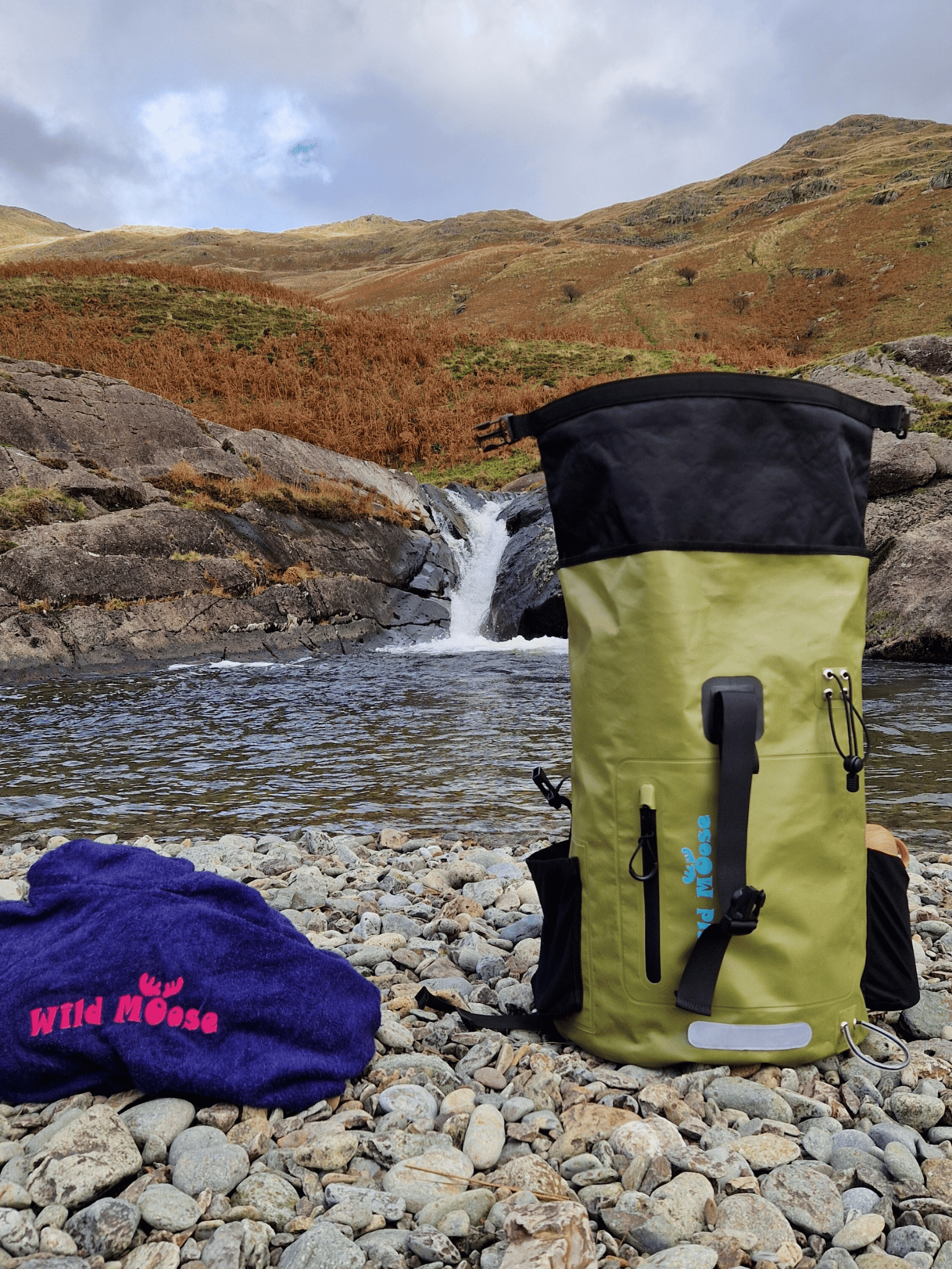 a green backpack and a blue Wild moose poncho towel on a pebble beach in front of a small waterfall pool with mountains in the background
